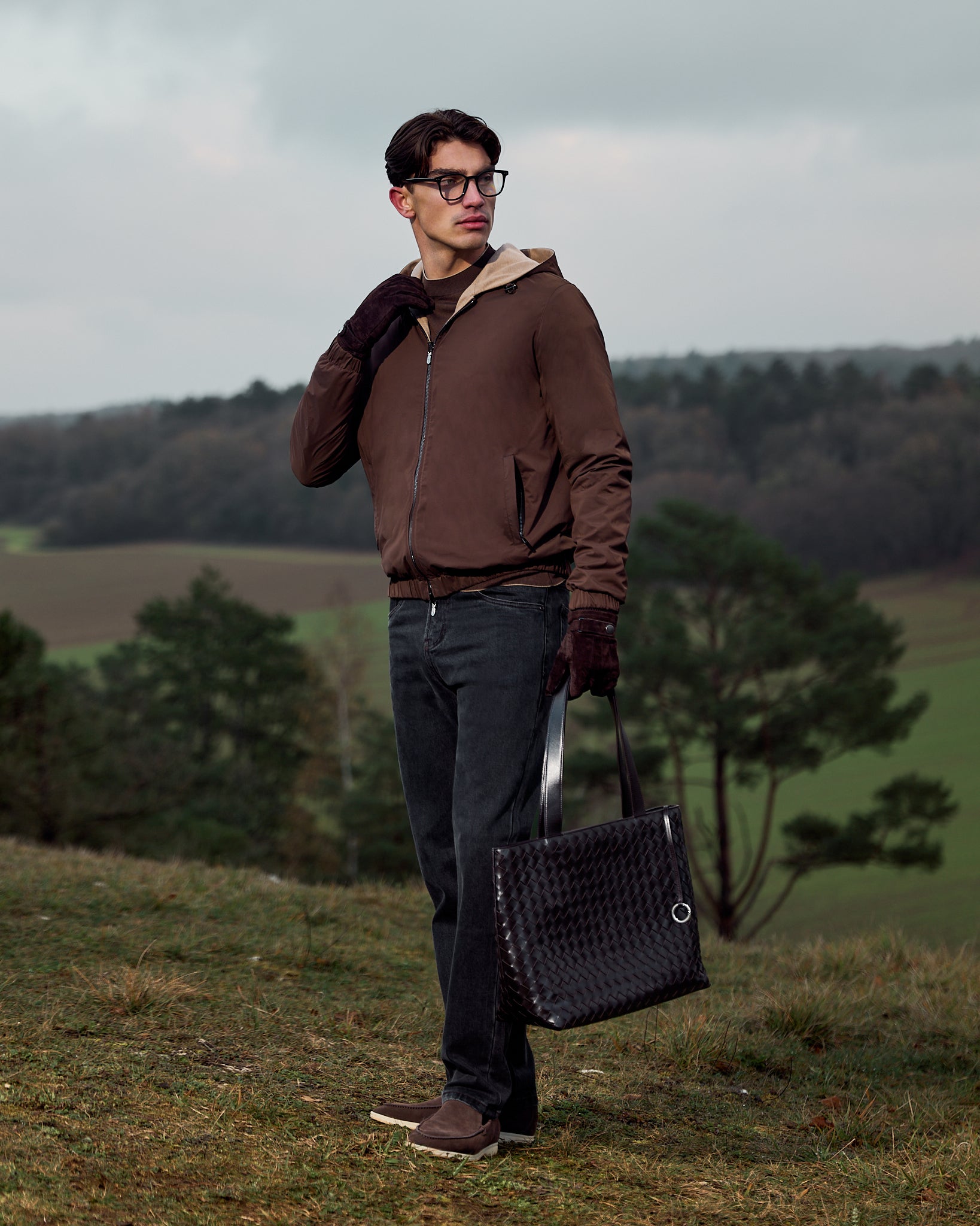 Man in brown jacket and dark pants holding a black bag in a field