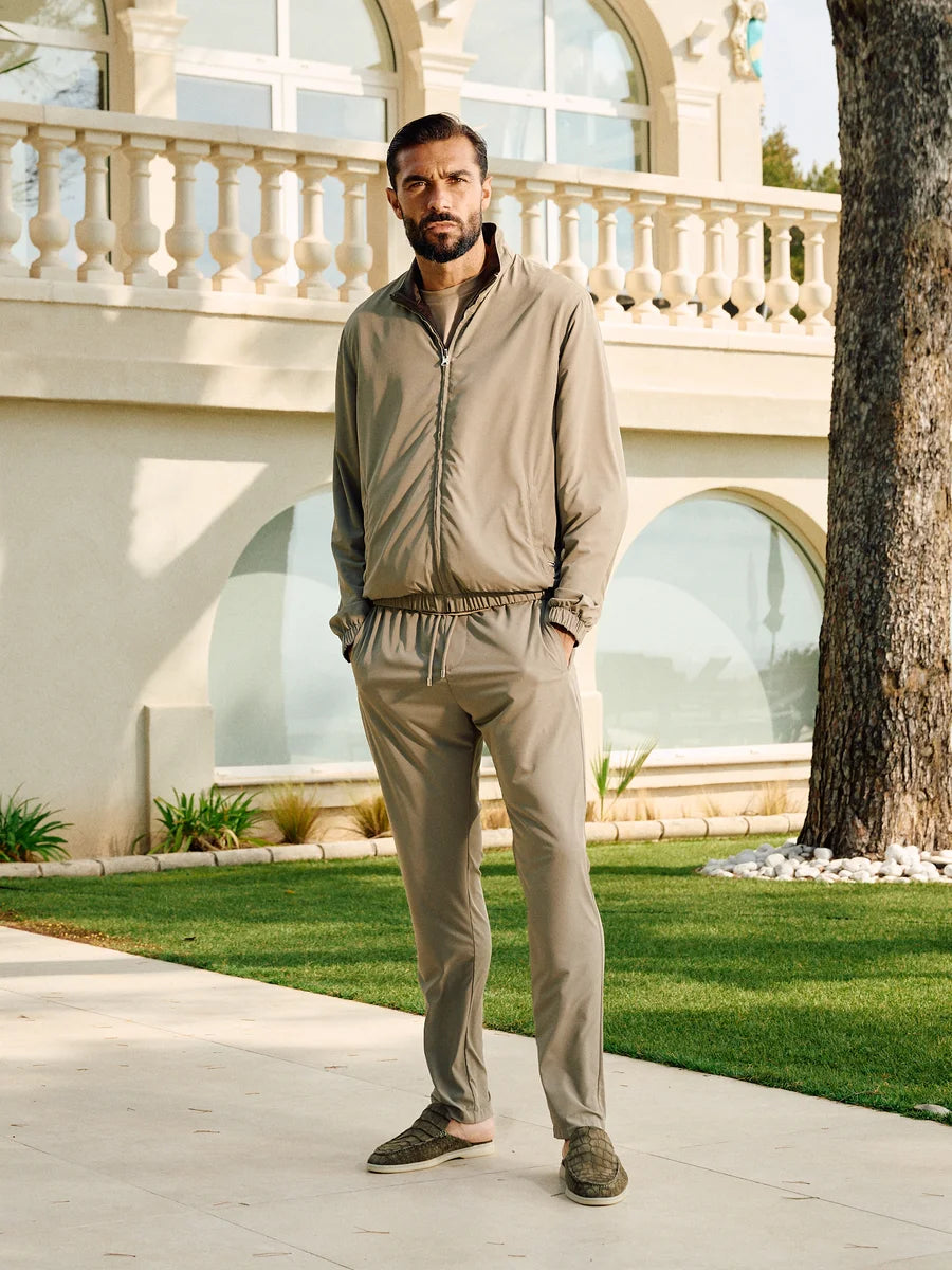 Man in beige tracksuit standing in front of a luxurious building with a fountain.