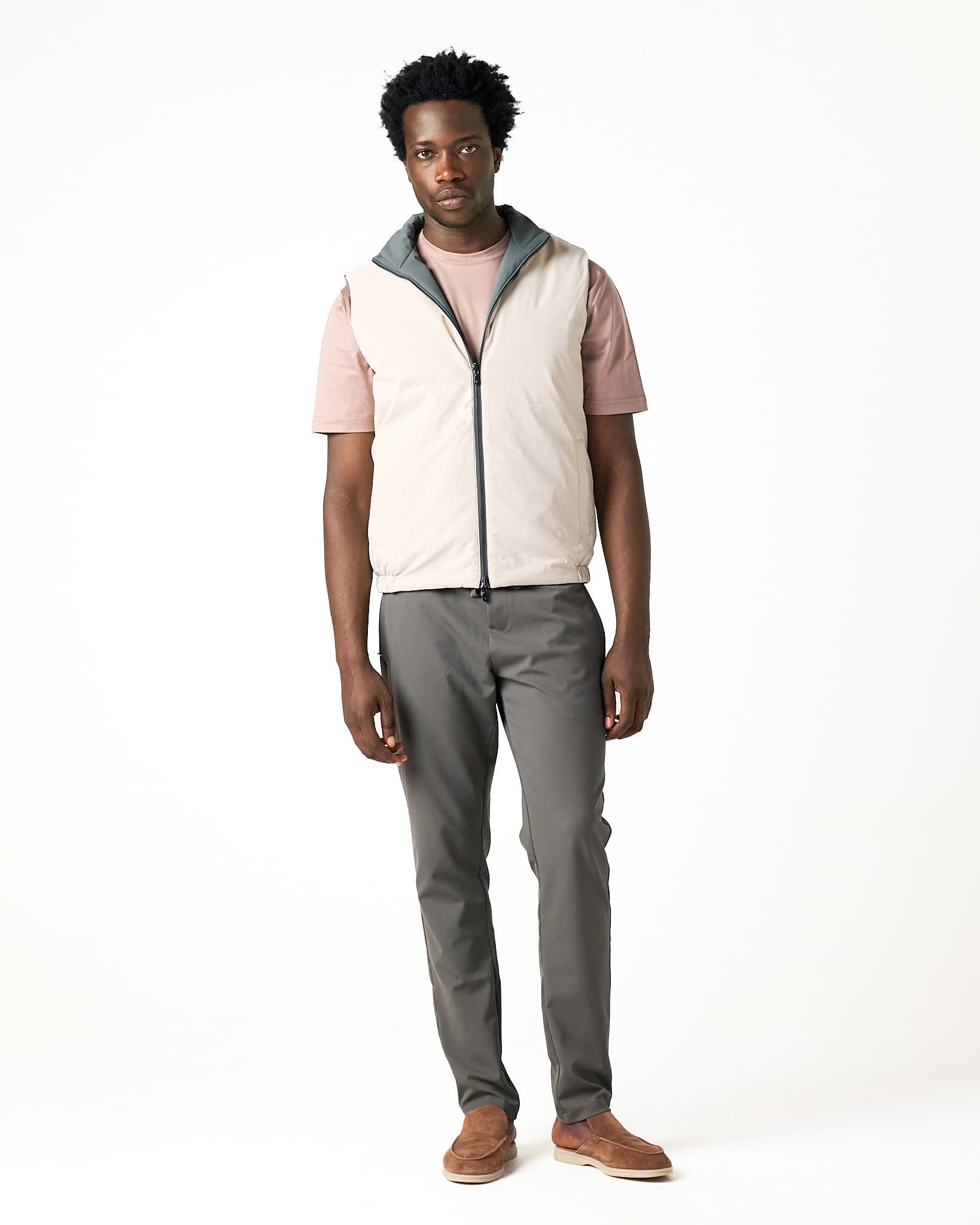 Man wearing a light-colored vest over a pink shirt, gray pants, and brown shoes on a white background