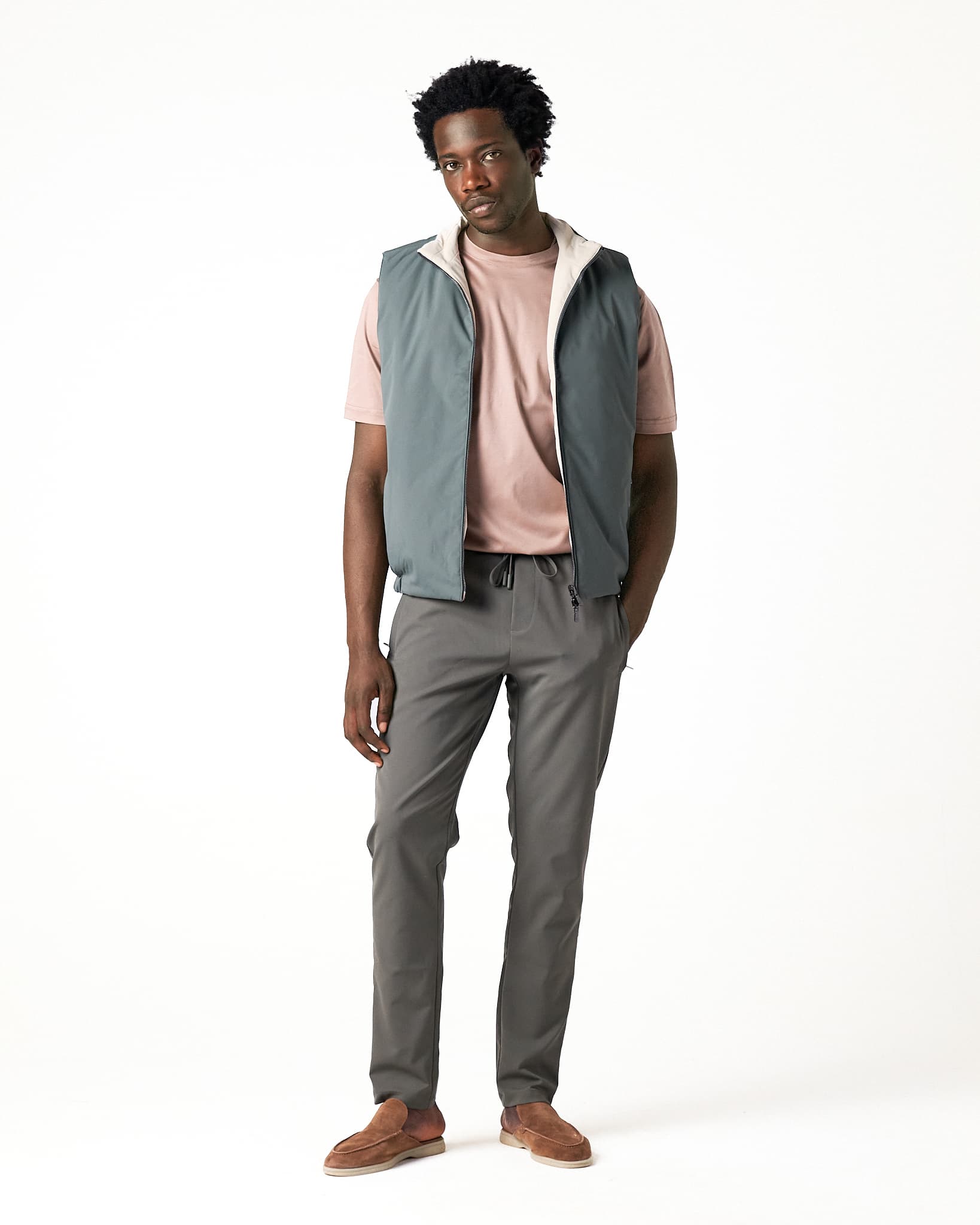 Man wearing a light gray vest over a pink shirt with gray pants on a white background