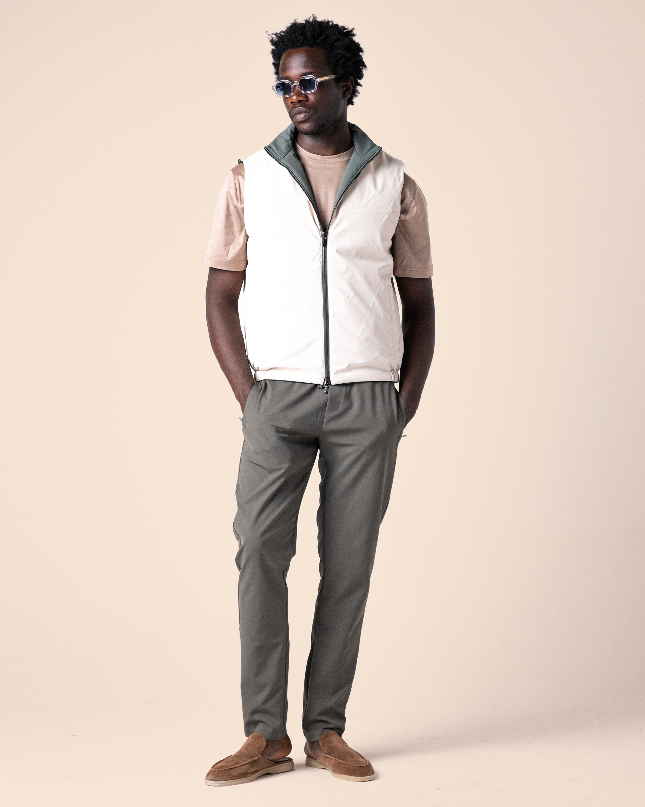 Man wearing a white vest over a beige shirt with gray pants on a beige background