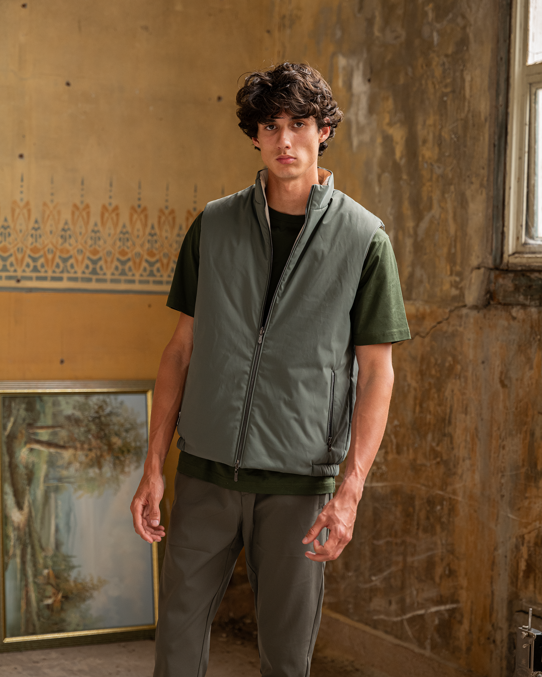 Man wearing a green vest over a black shirt in a room with peeling paint and a painting on the wall.