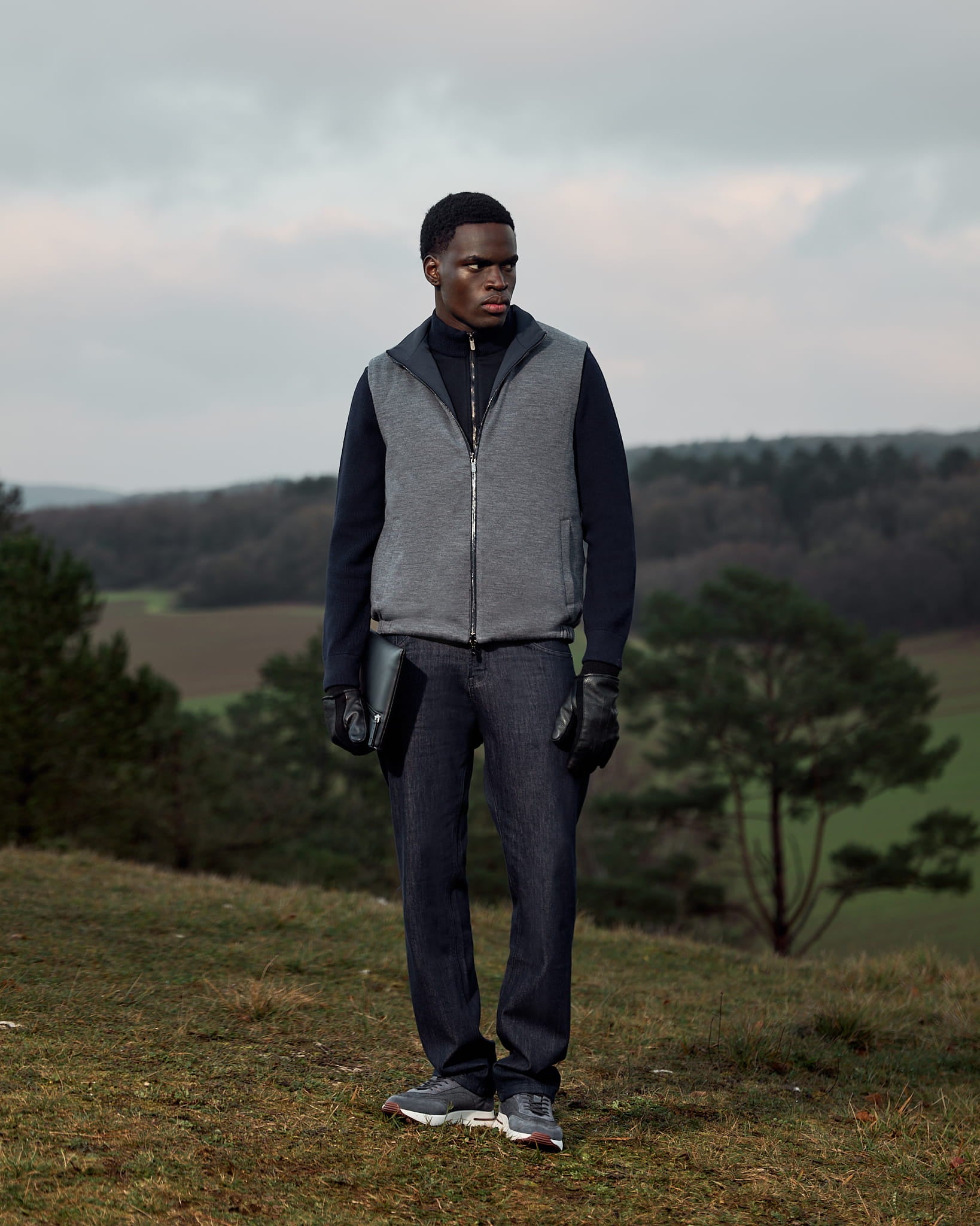 Person wearing a gray vest and dark pants standing in a field with trees and a cloudy sky.