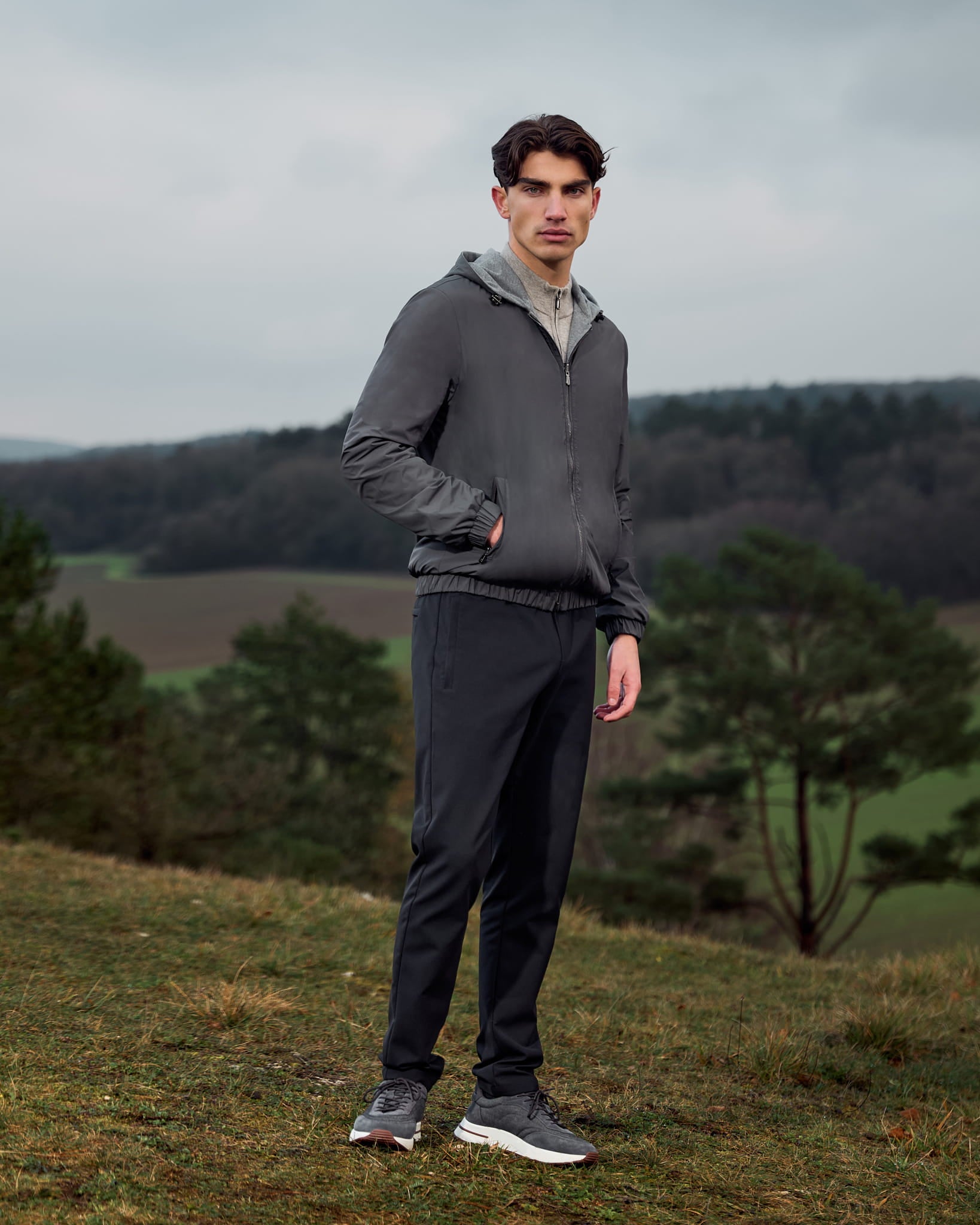 Man wearing a gray jacket and black pants standing in a natural landscape with trees and open fields.