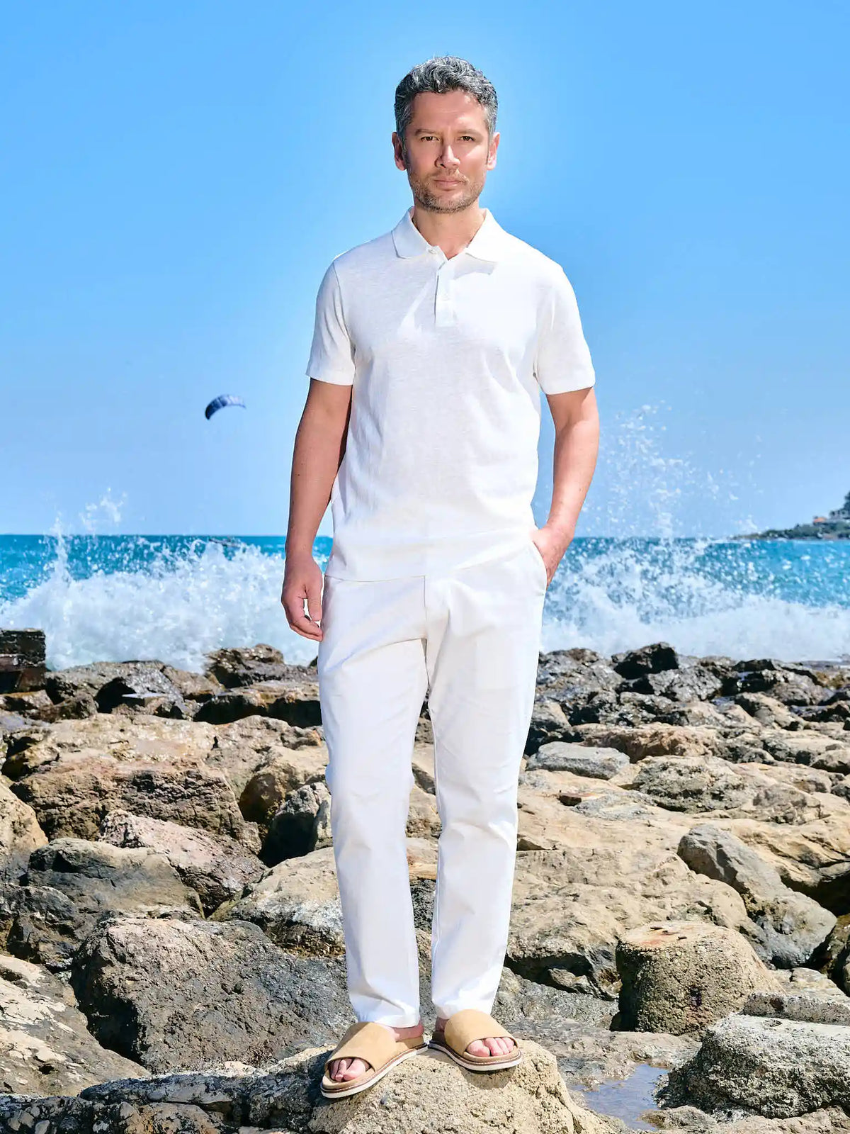 Man in an ivory polo and white trousers on a rocky shore