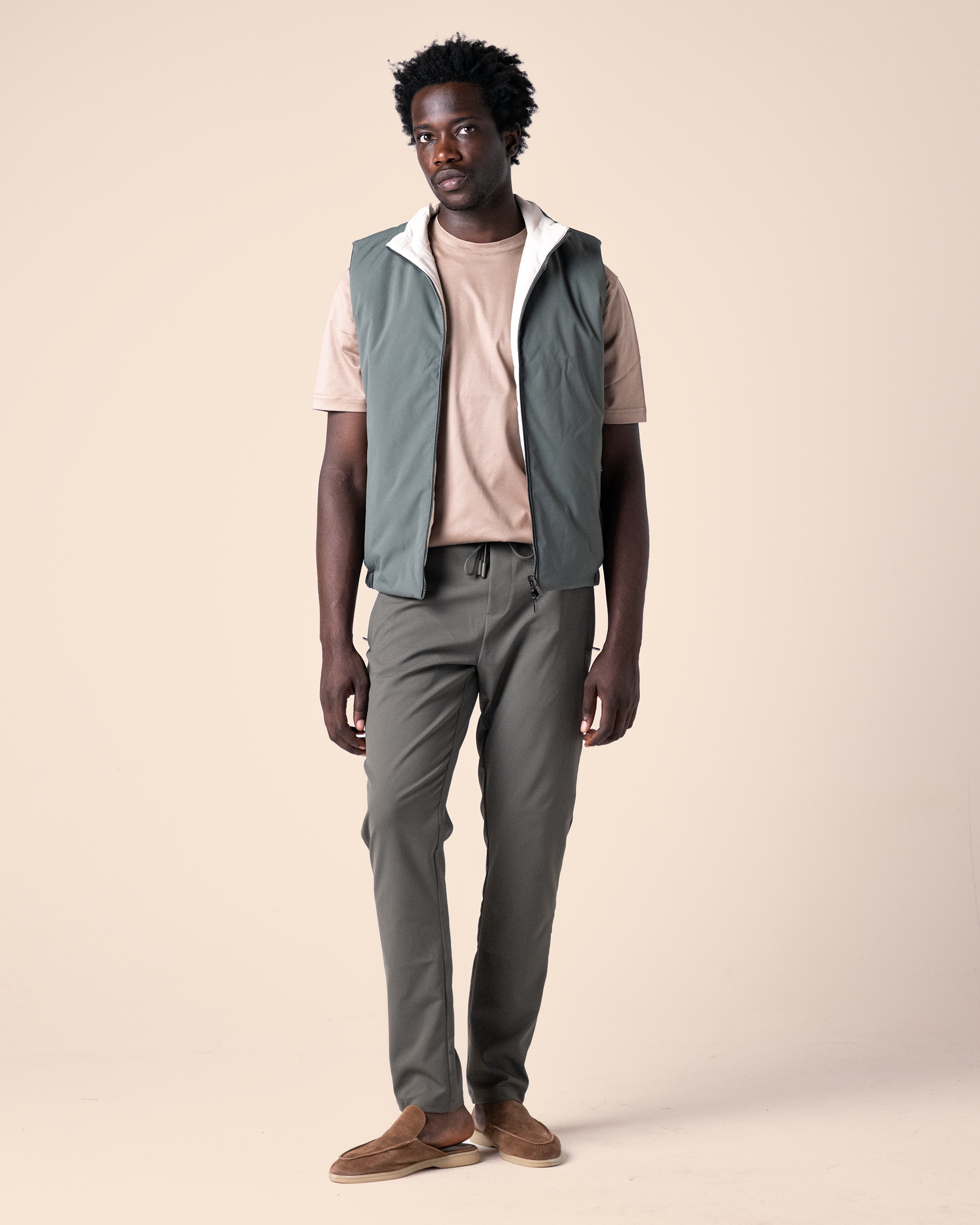 Man wearing a light gray vest over a beige shirt with dark pants on a beige background