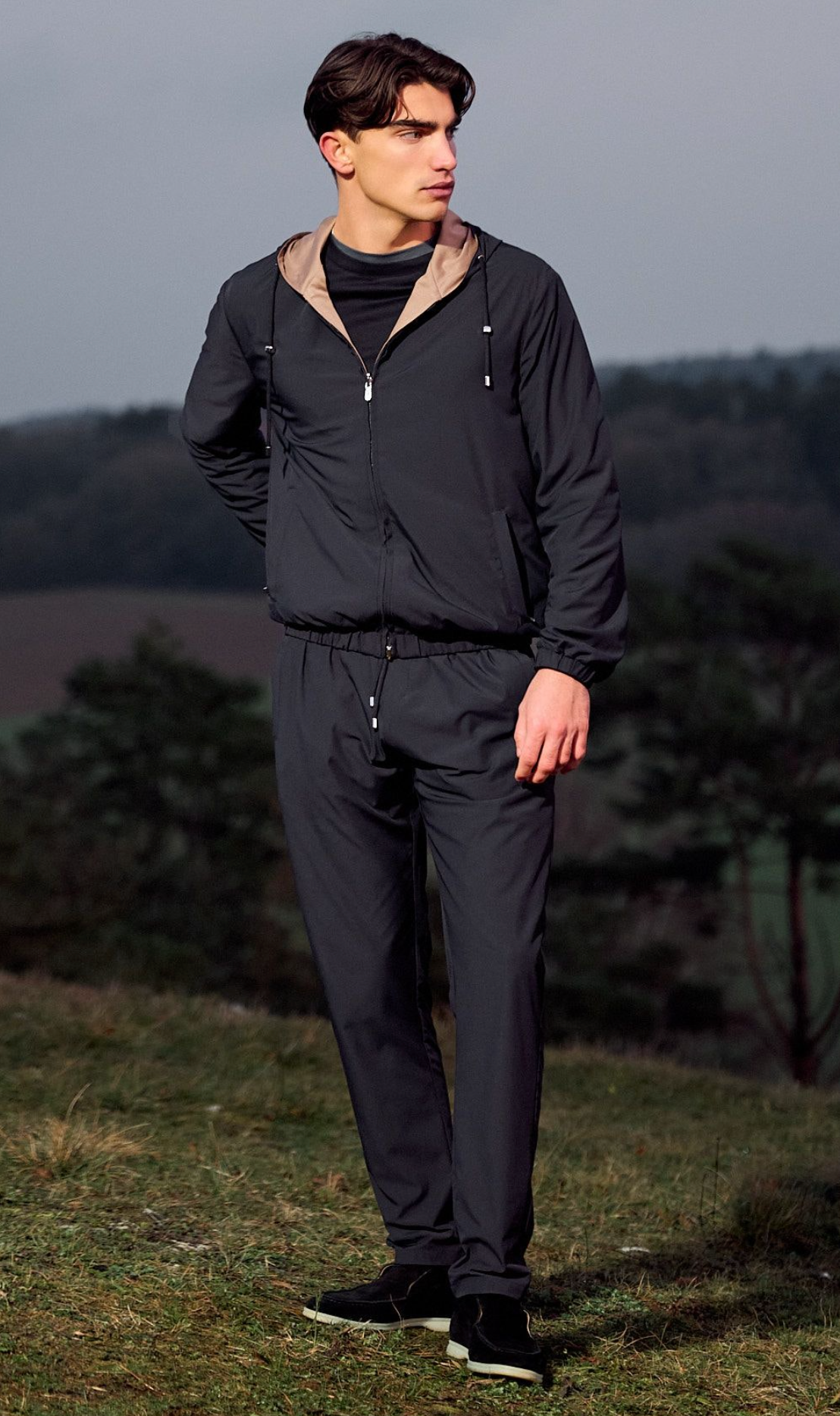 Man wearing a dark jacket and pants standing outdoors with trees in the background