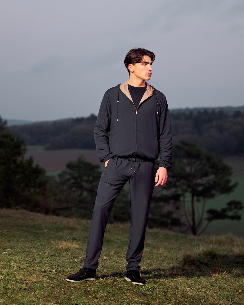 Man wearing a dark gray tracksuit standing outdoors with a landscape background