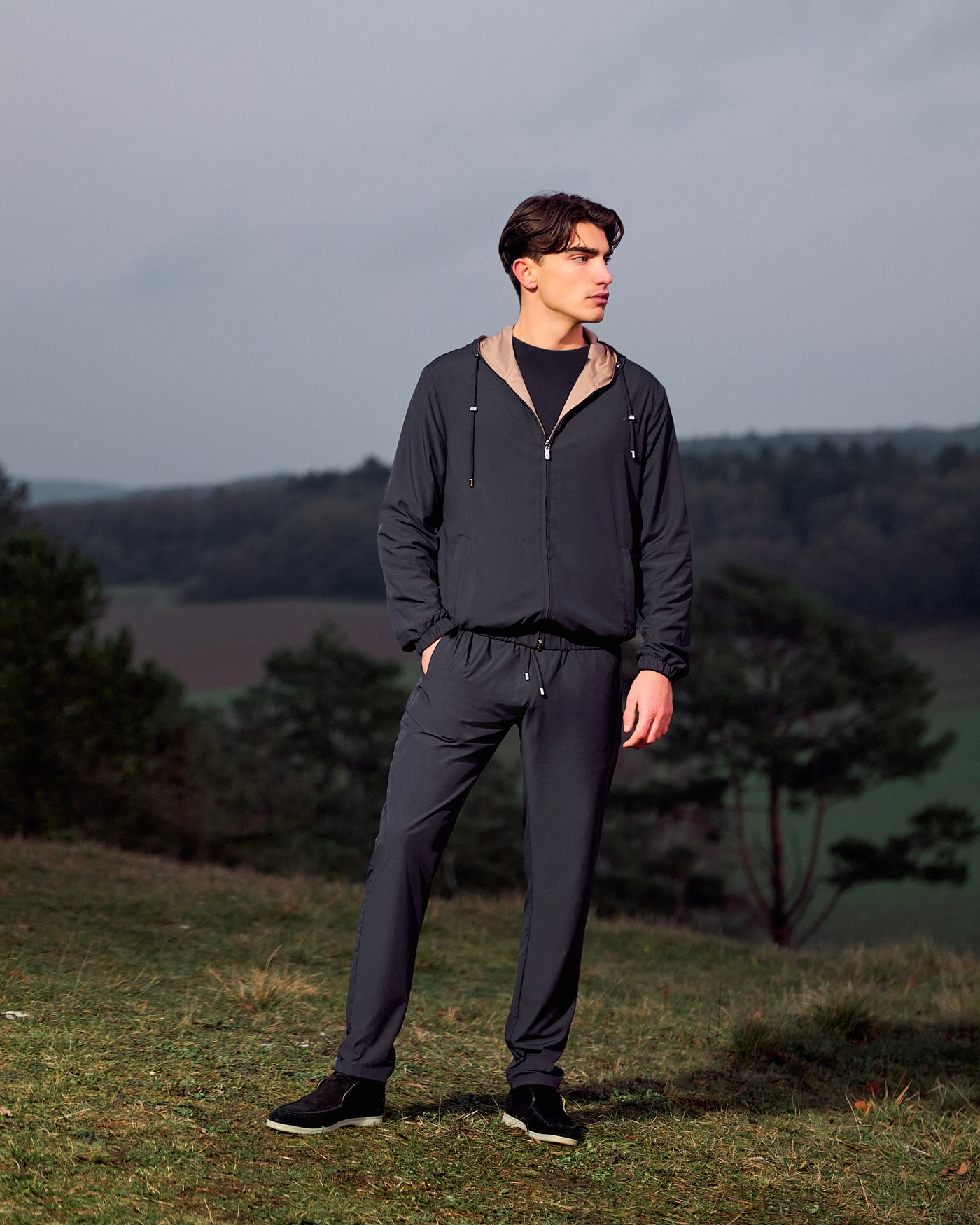 Man wearing a dark gray tracksuit standing outdoors with a landscape background