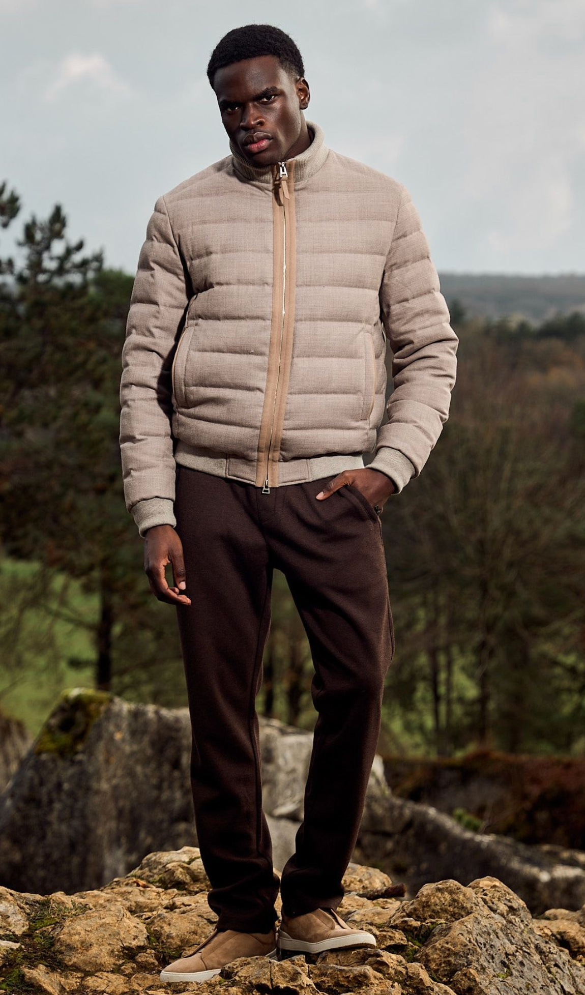 Man wearing a beige puffer jacket and brown pants standing on rocky terrain with trees in the background