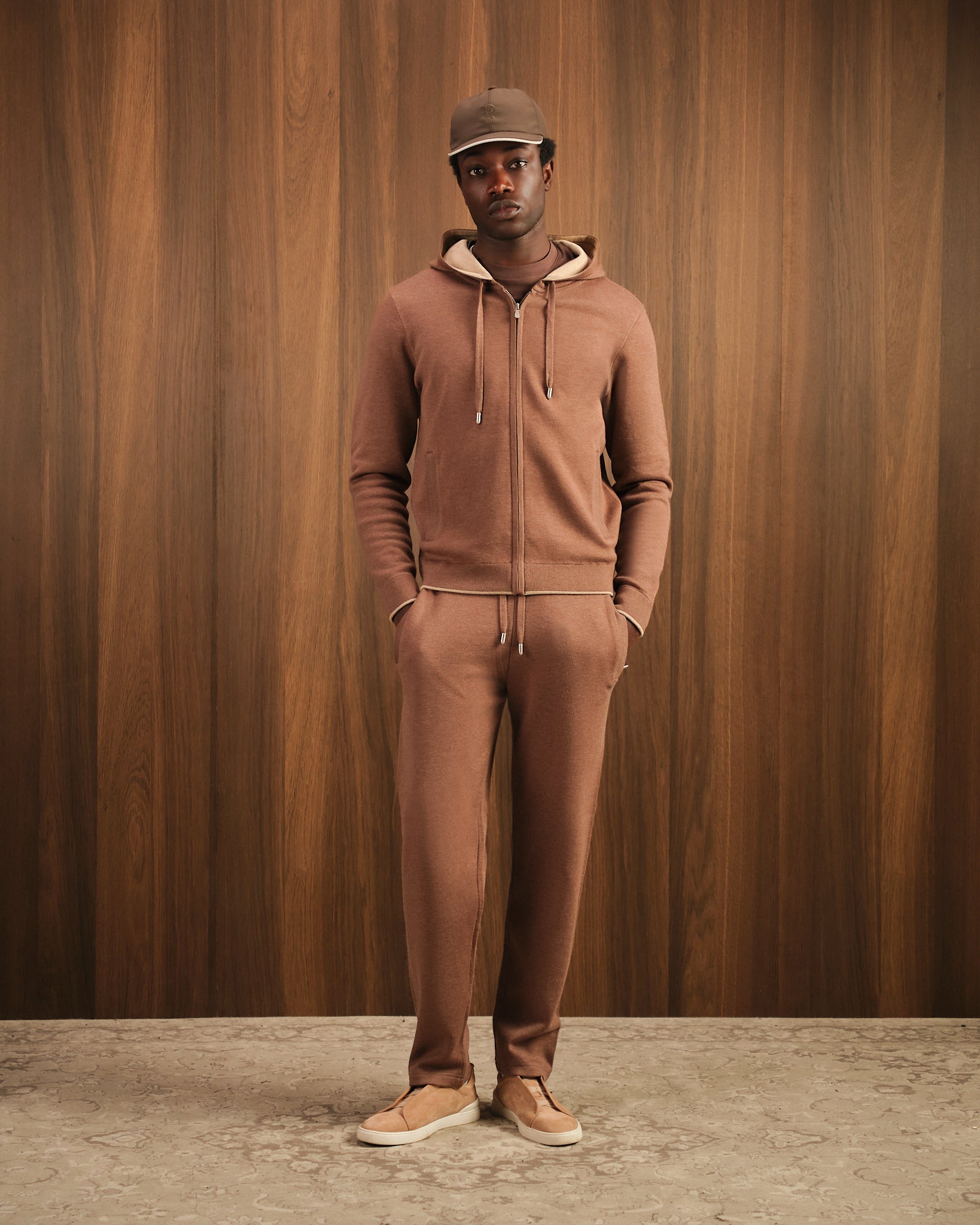 Man wearing a brown tracksuit and cap against a wooden paneled wall