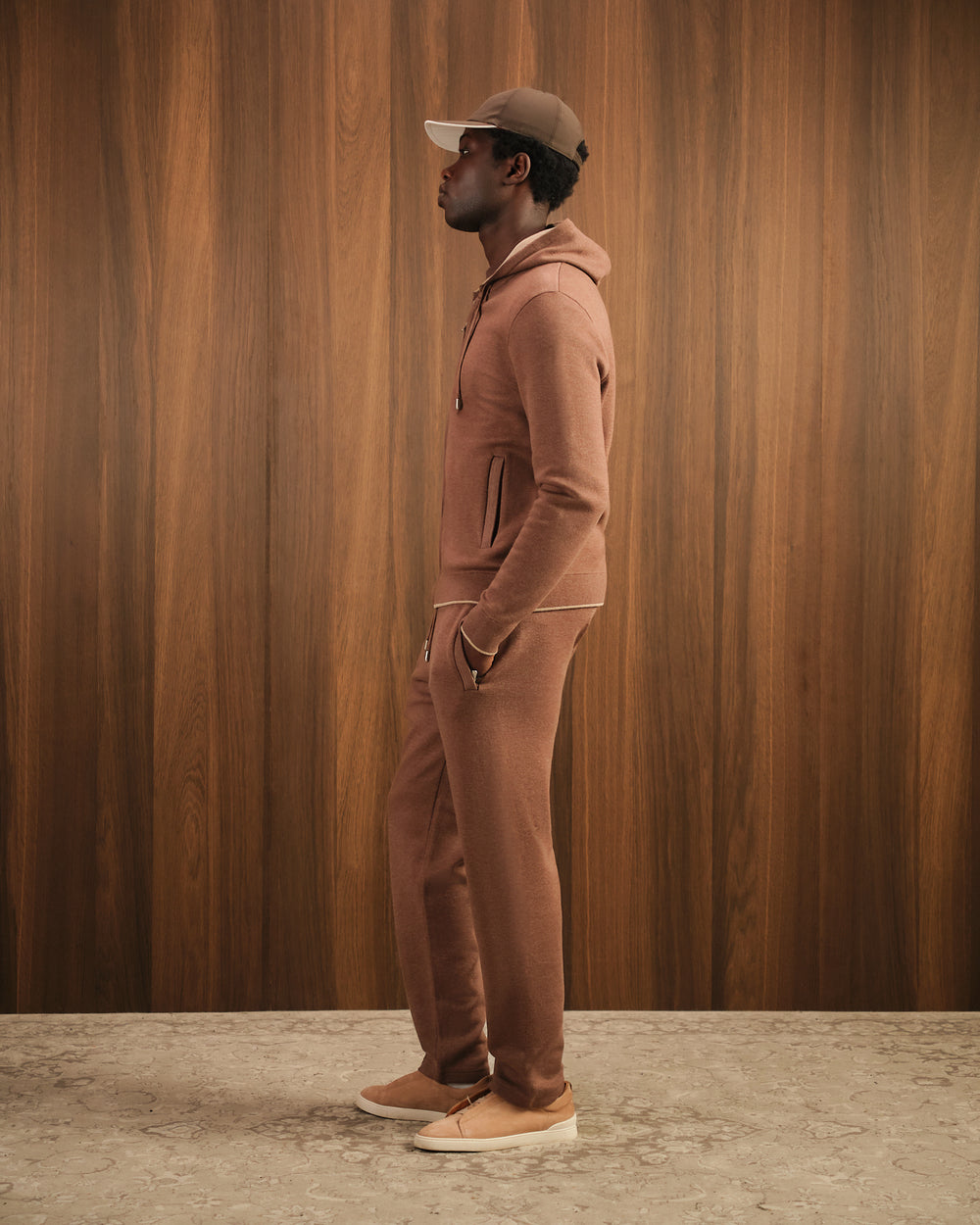 Man wearing a brown tracksuit and cap against a wooden paneled wall.