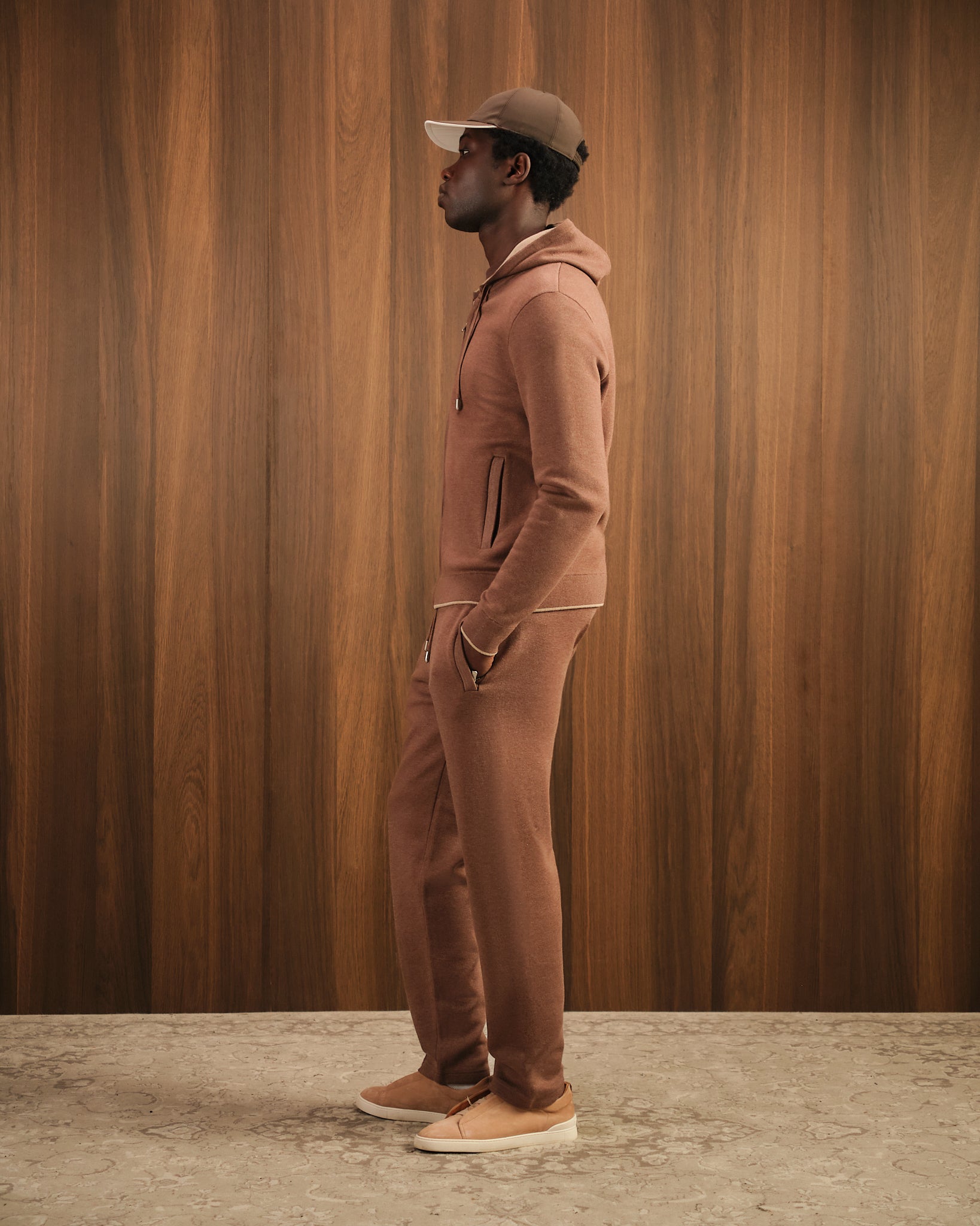 Man wearing a brown tracksuit and cap against a wooden paneled wall.