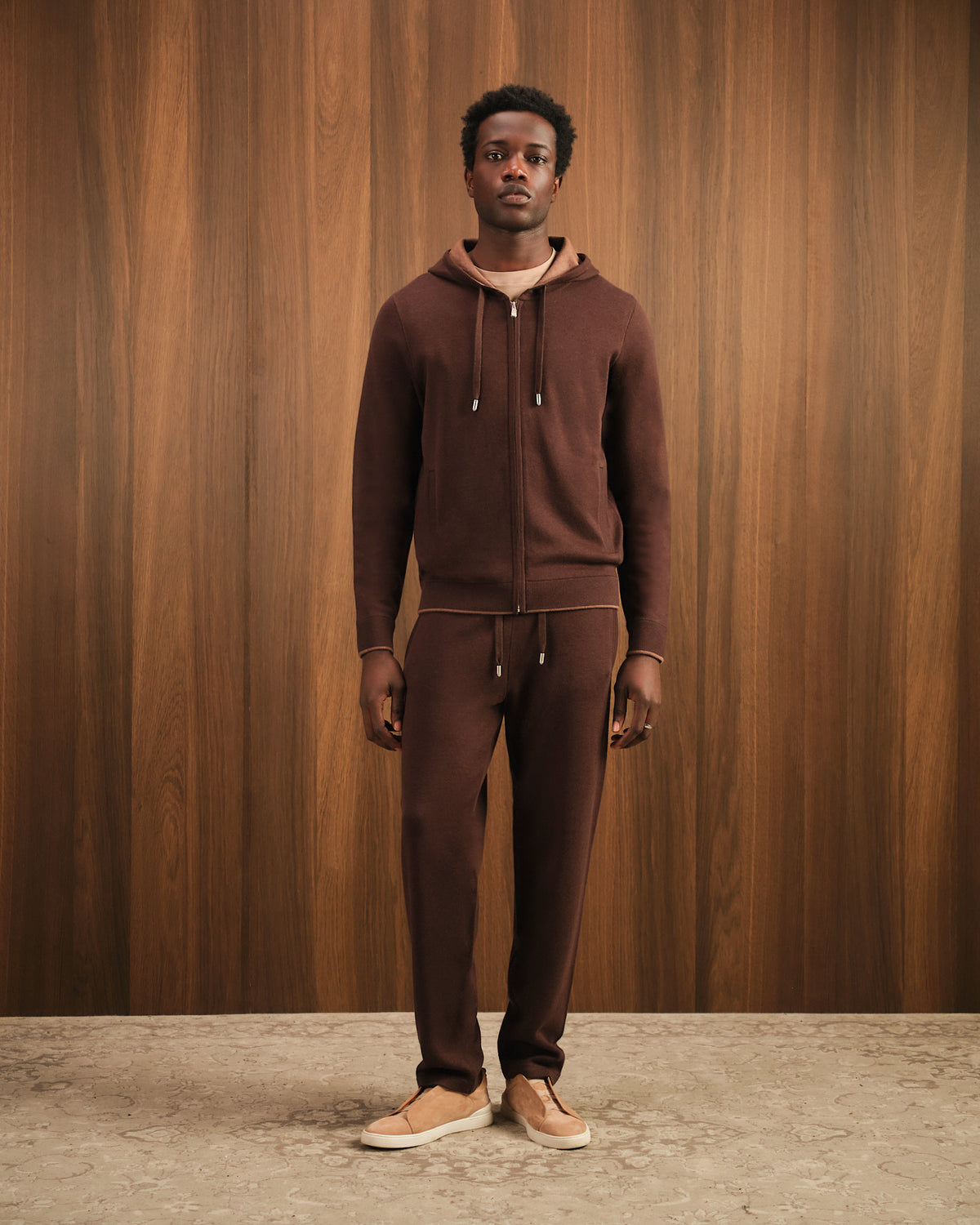 Man wearing a brown tracksuit standing against a wooden paneled wall.