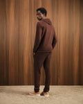 Man wearing a brown tracksuit standing against a wooden paneled wall.