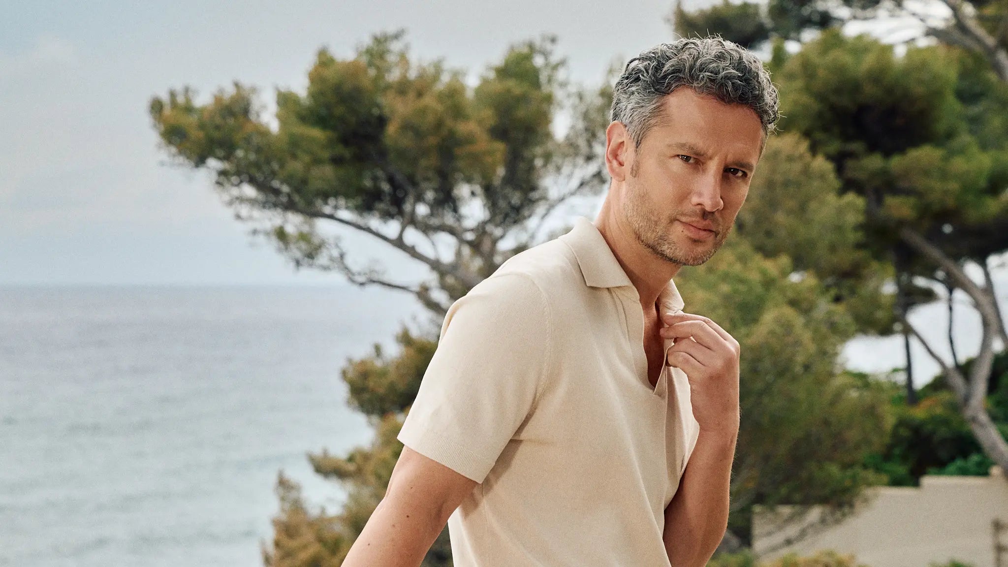 Man in a beige shirt standing by trees with a body of water in the background
