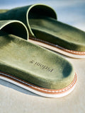 Close-up of olive green slide sandals with a suede-like finish, white stitching, and a tan outsole on a light neutral surface. 