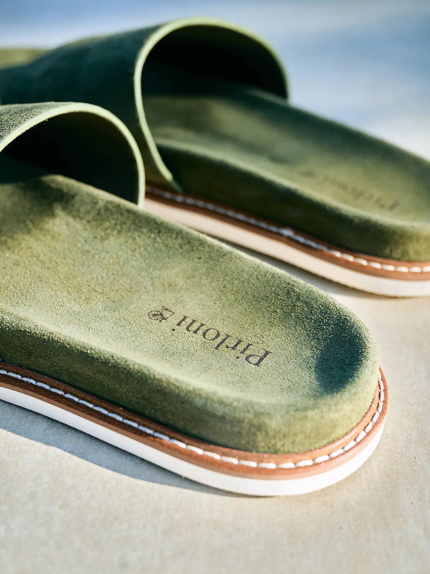 Close-up of olive green slide sandals with a suede-like finish, white stitching, and a tan outsole on a light neutral surface. 