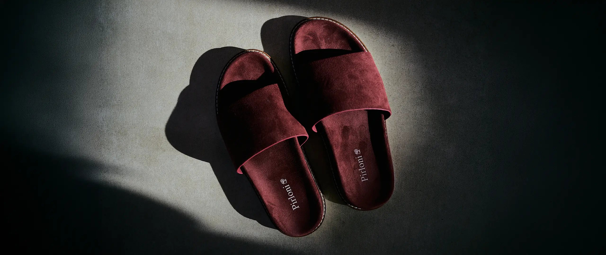 A pair of deep burgundy suede slide sandals with a thin brown sole, displayed on a dark gray textured surface under dramatic, directional sunlight that casts a strong shadow. The brand name 'Pimkie' is printed on the insole.