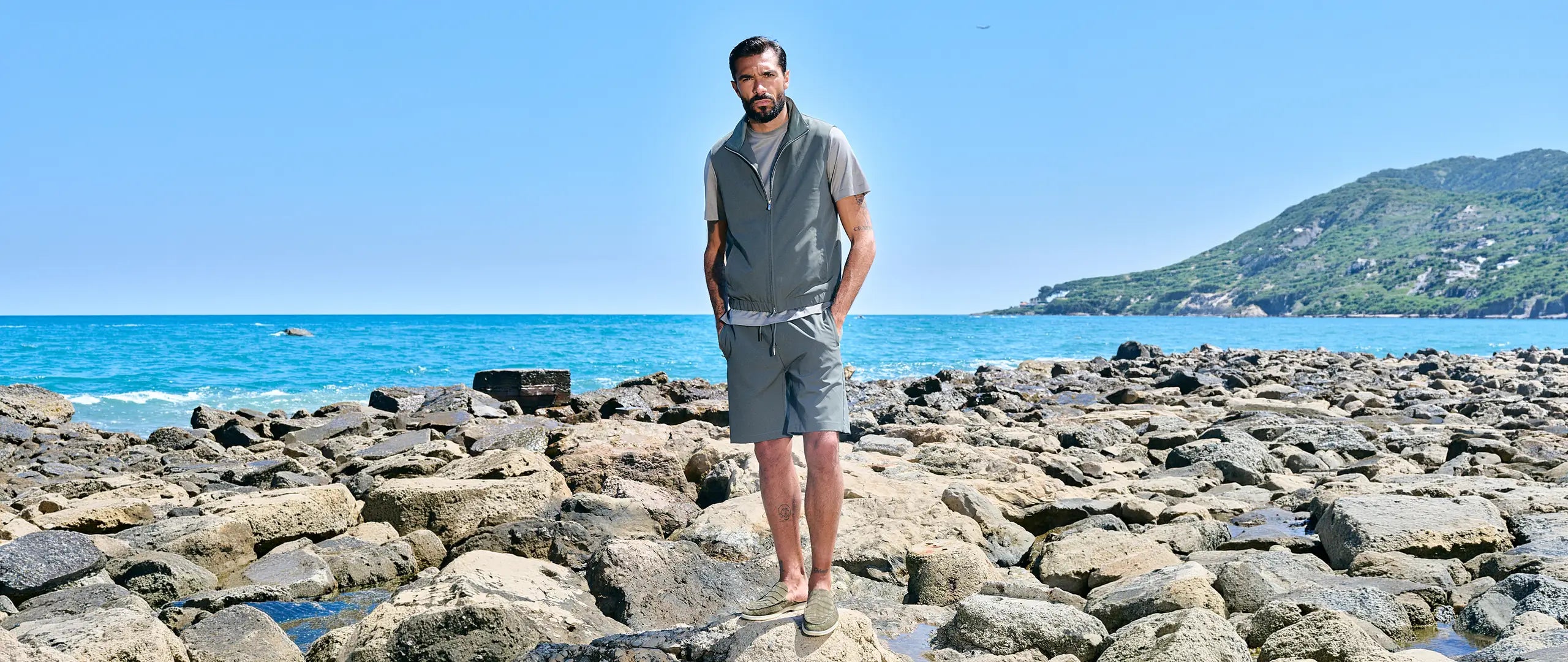A man wearing a gray sleeveless zip-up vest and matching gray shorts stands on a rocky beach with the blue sea and mountains in the background.