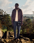 Man wearing a brown puffer jacket standing on rocky terrain with trees and mountains in the background