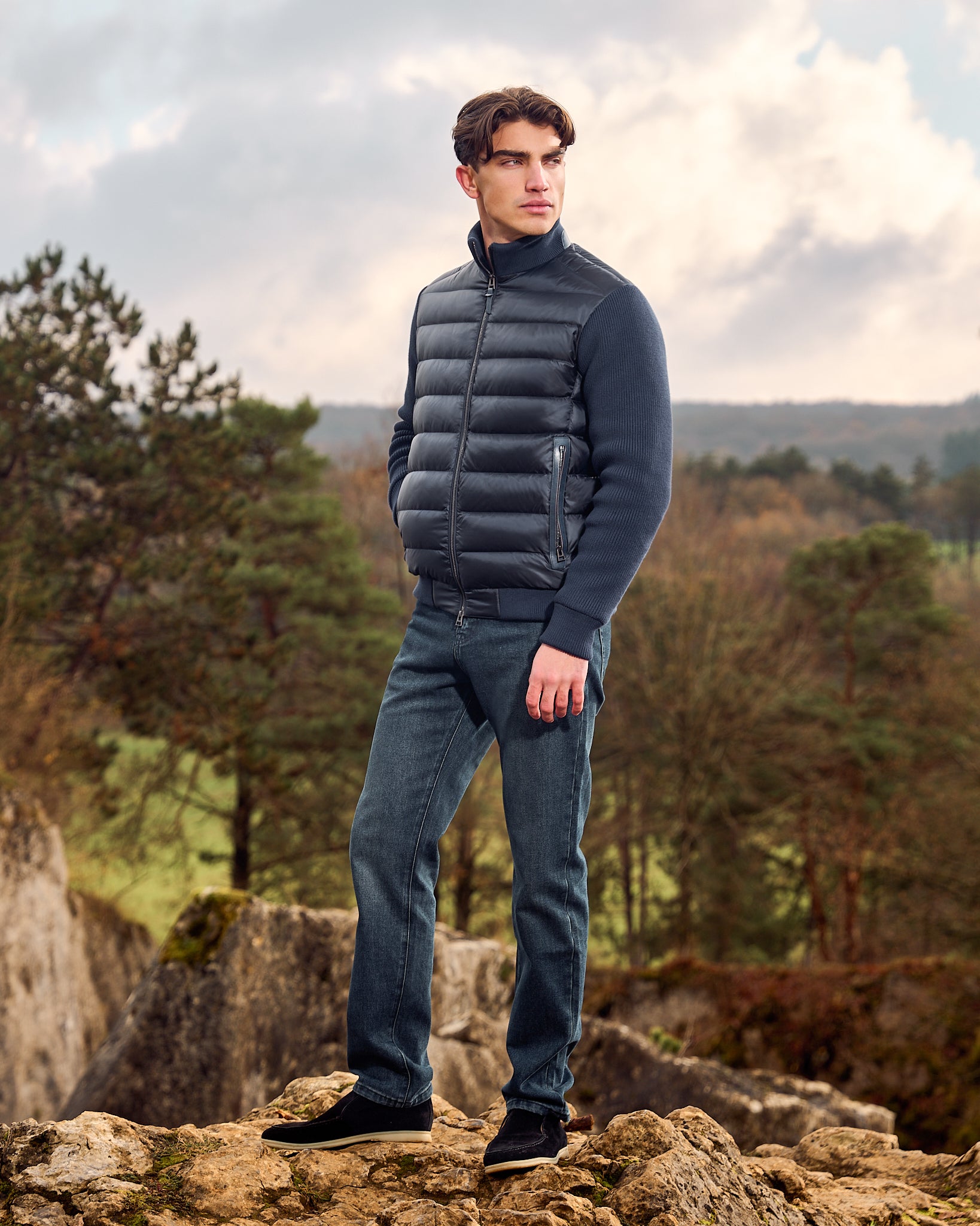 Man wearing a dark blue puffer jacket and jeans standing on rocky terrain with trees and sky in the background