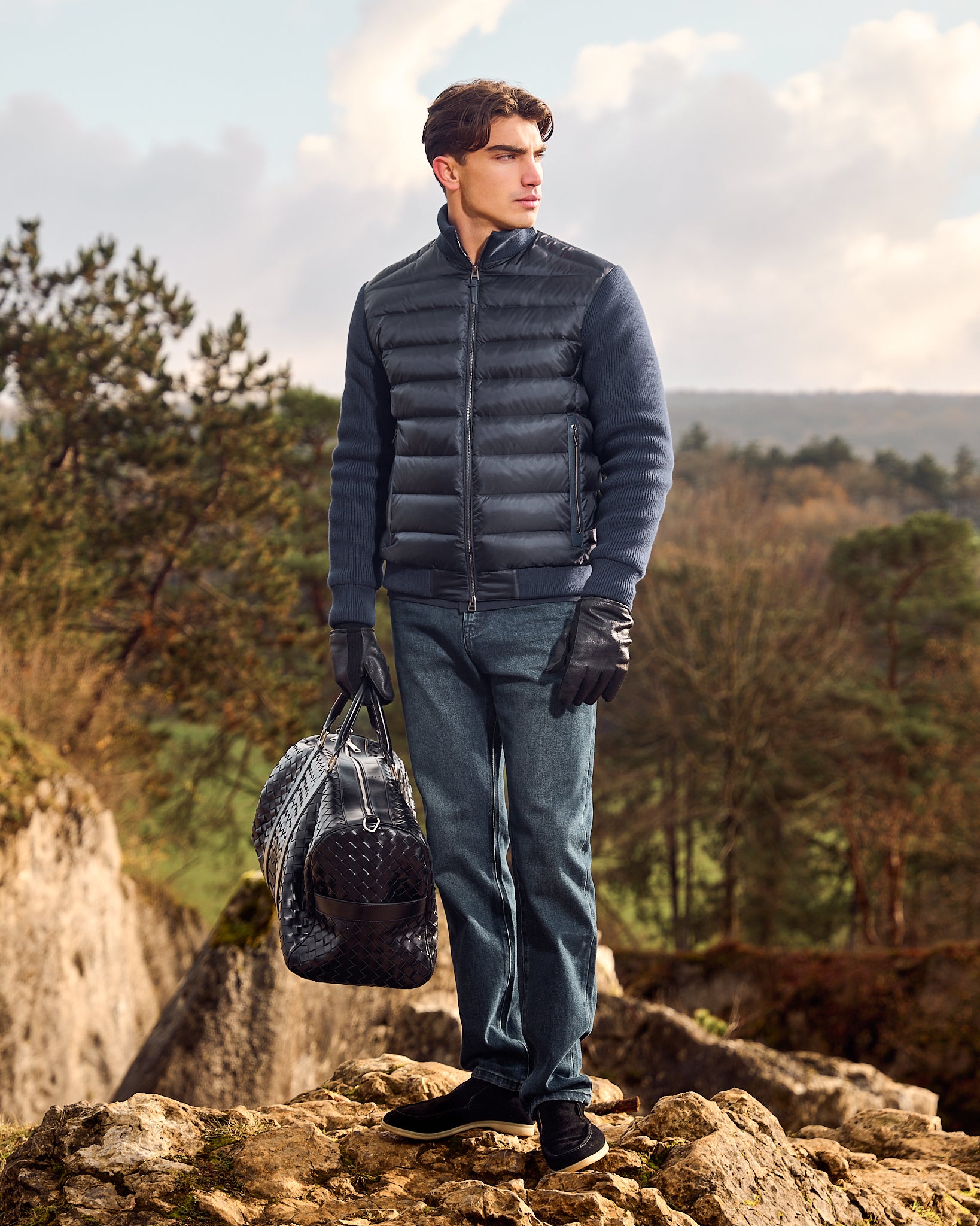 Man in a blue puffer jacket holding a bag on a rocky landscape with trees and sky.