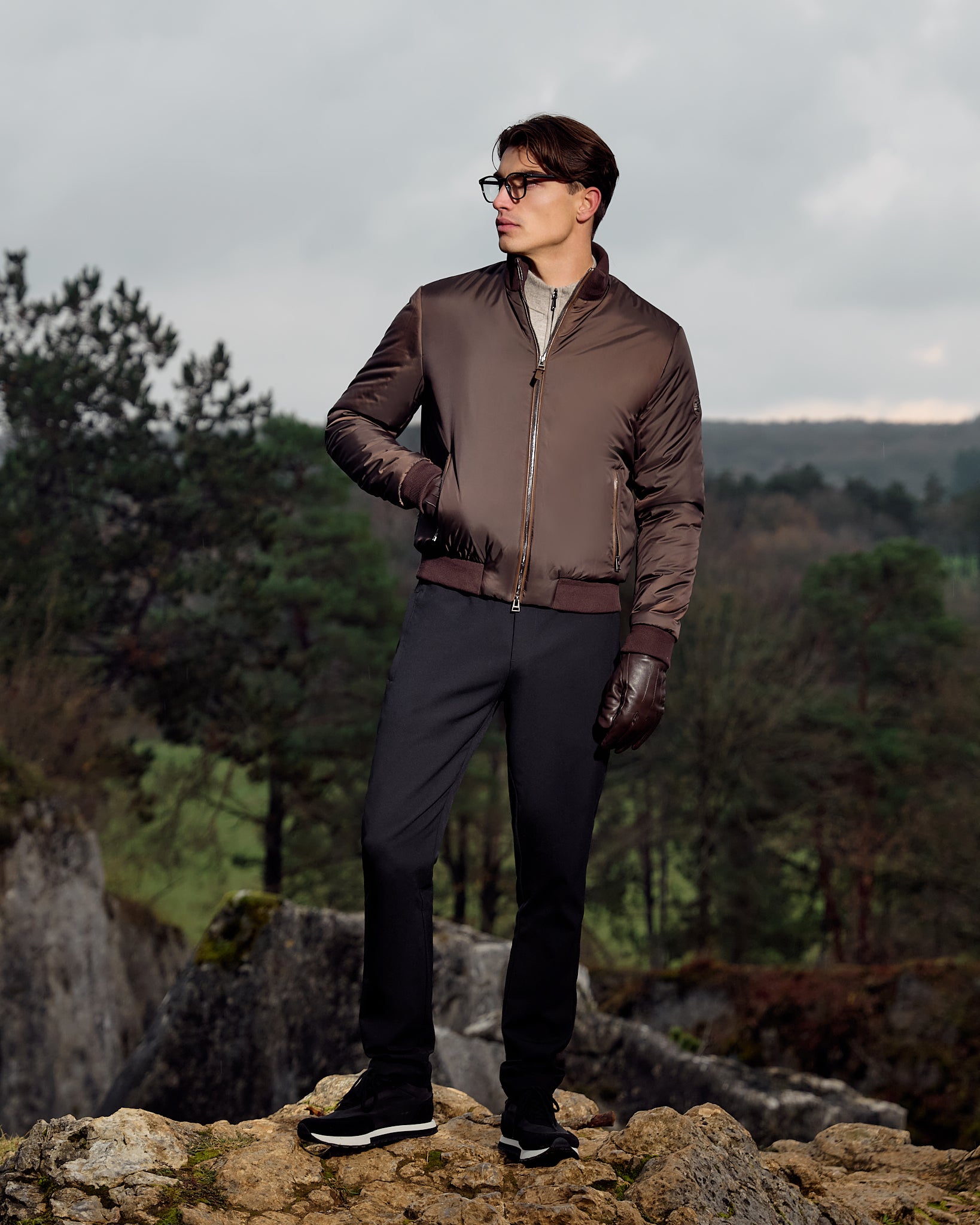 Man wearing a brown jacket and black pants standing on a rocky outcrop with a forest background