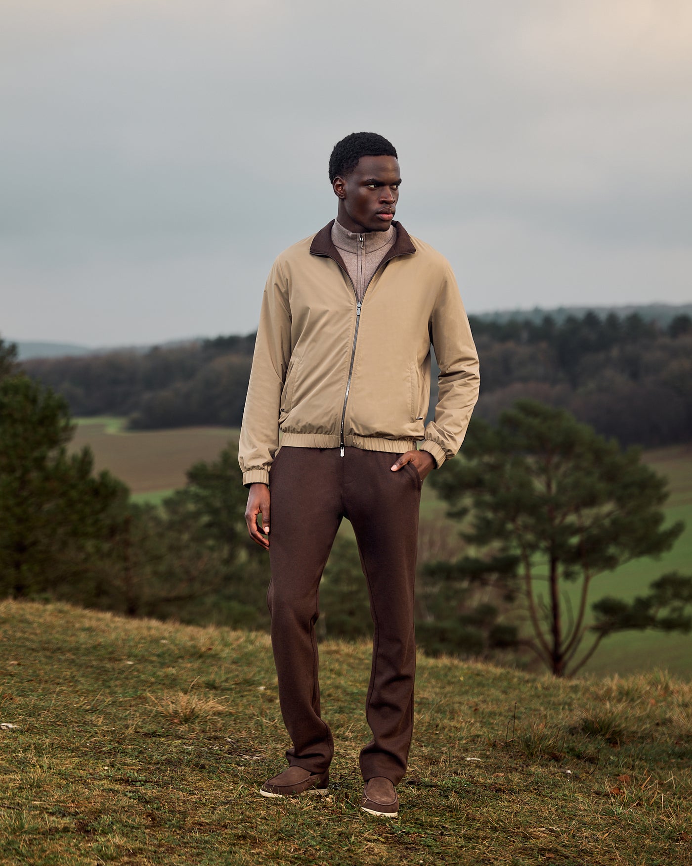 Man standing in a field wearing a beige jacket and brown pants.