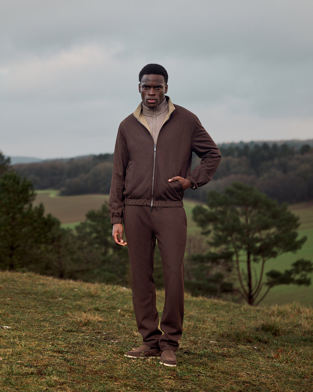 Man in a brown tracksuit standing in a field with trees and a cloudy sky in the background