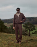 Man in a brown tracksuit standing in a field with trees and a cloudy sky in the background