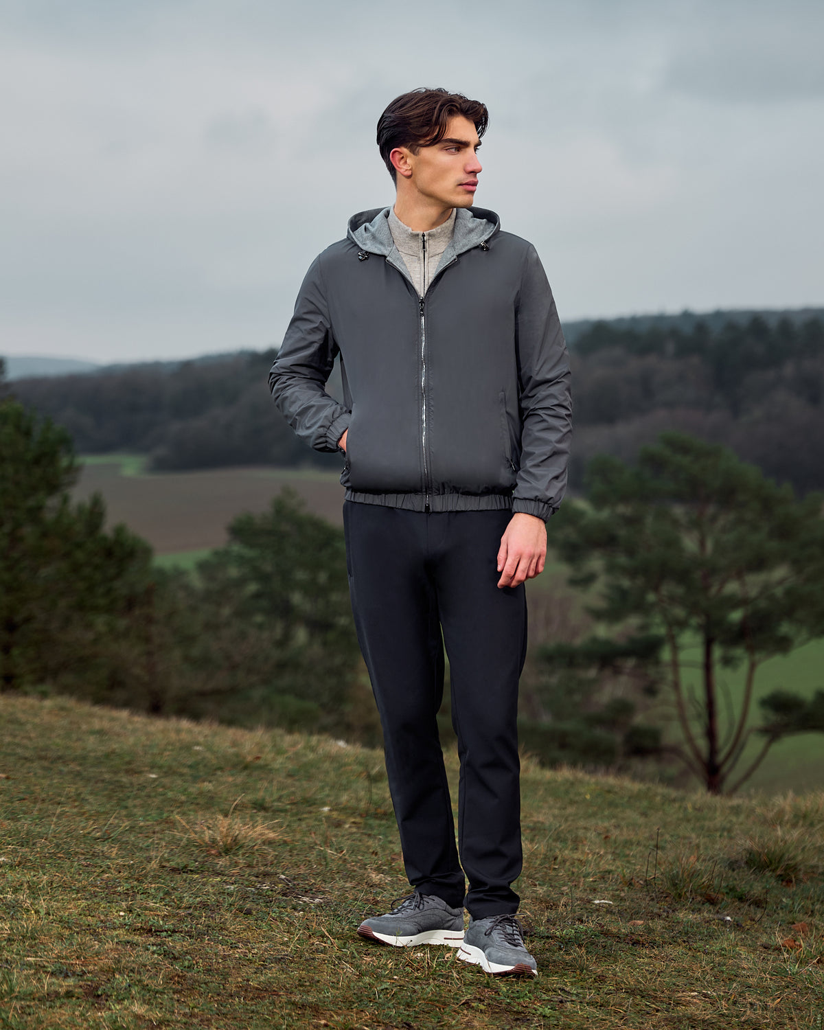 Man wearing a gray jacket and black pants standing outdoors with trees and fields in the background