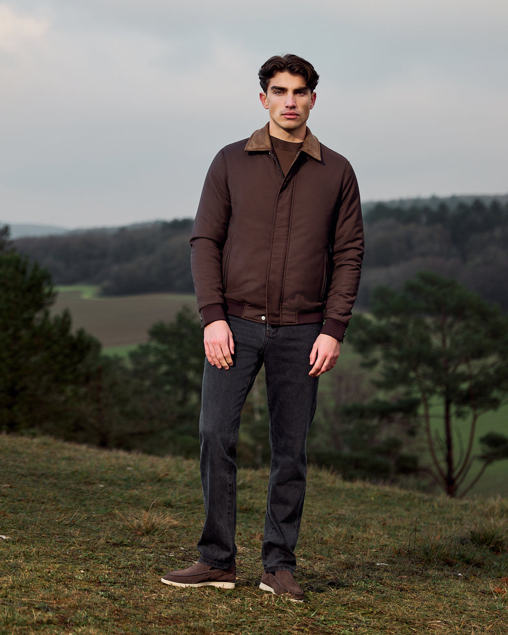 Man wearing a brown jacket and dark pants standing in a natural landscape.