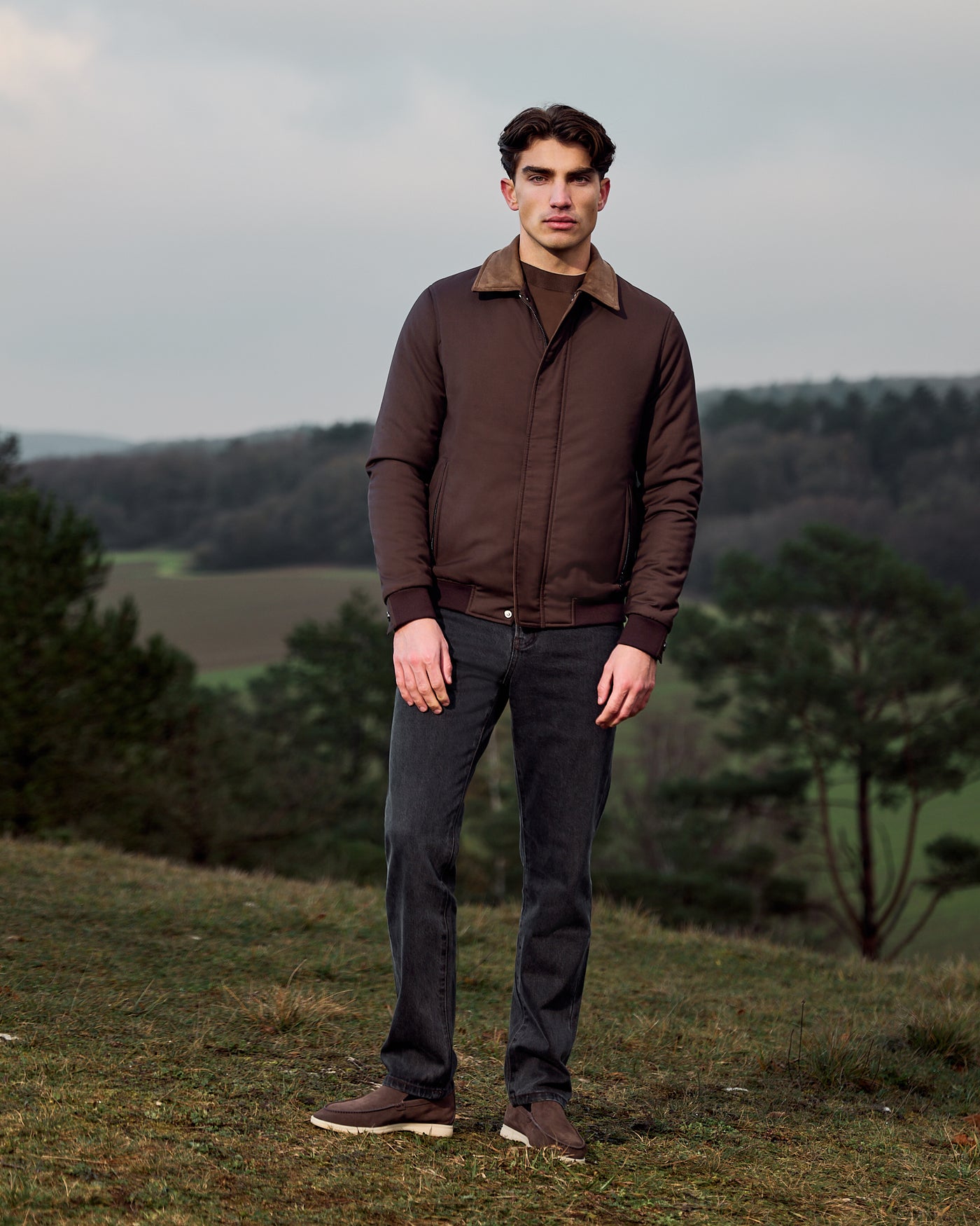 Man wearing a brown jacket and dark pants standing in a natural landscape.