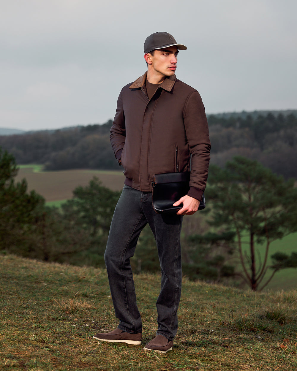 Man wearing a brown jacket and cap standing outdoors with trees and fields in the background