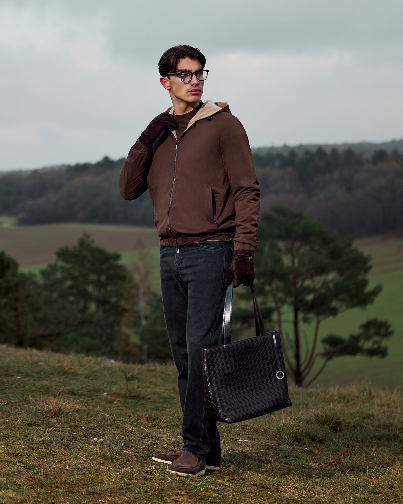 Man in brown jacket and dark pants holding a black bag in a field