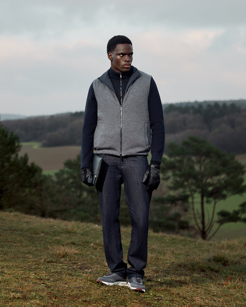 Man wearing a gray vest and dark pants standing in a field with trees and sky in the background