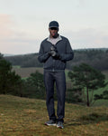 Man in outdoor clothing standing on a grassy hill with trees and sky in the background