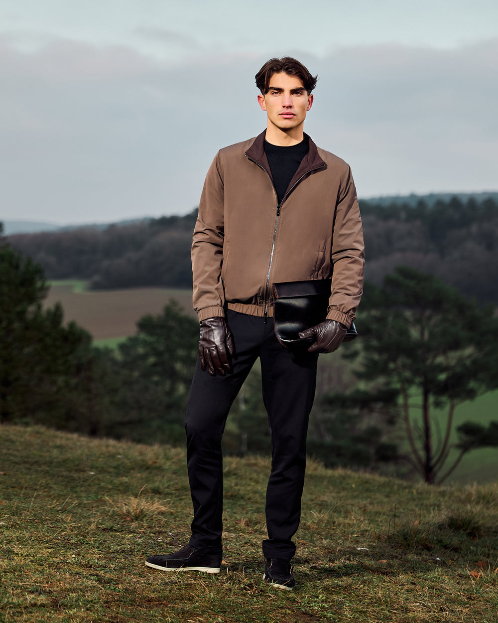 Man standing outdoors in a brown jacket and black pants holding a black helmet.