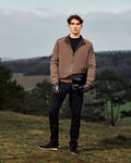 Man standing outdoors in a brown jacket and black pants holding a black helmet.