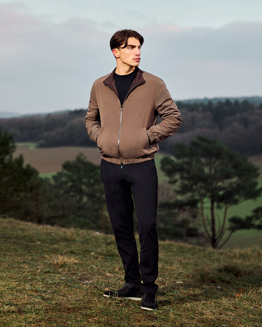 Man wearing a brown jacket standing in a natural landscape with trees and open fields.