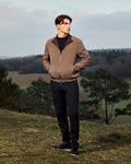 Man wearing a brown jacket standing in a natural landscape with trees and open fields.