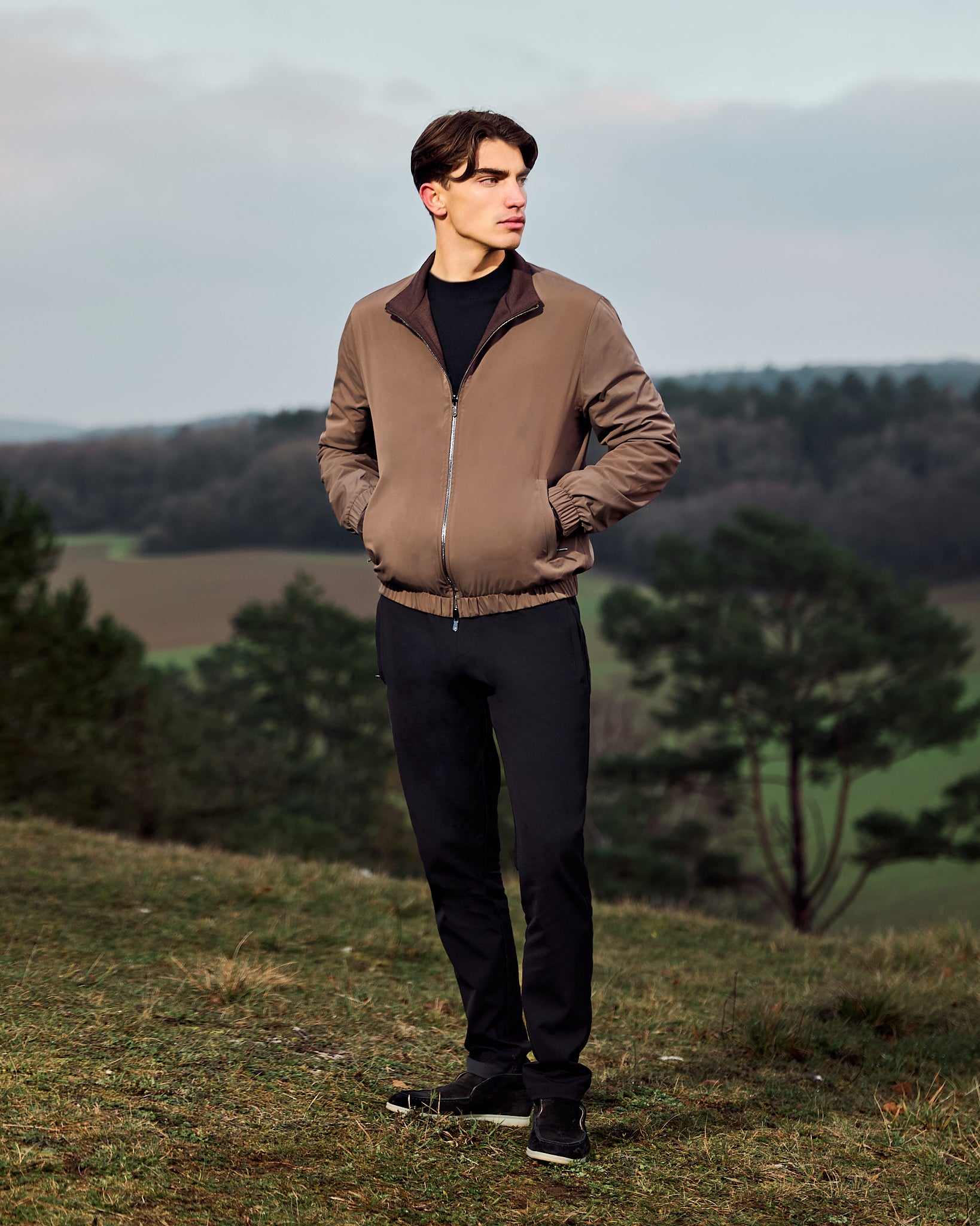 Man wearing a brown jacket standing in a natural landscape with trees and open fields.