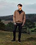 Man wearing a brown jacket and black pants standing in a field with trees and sky in the background