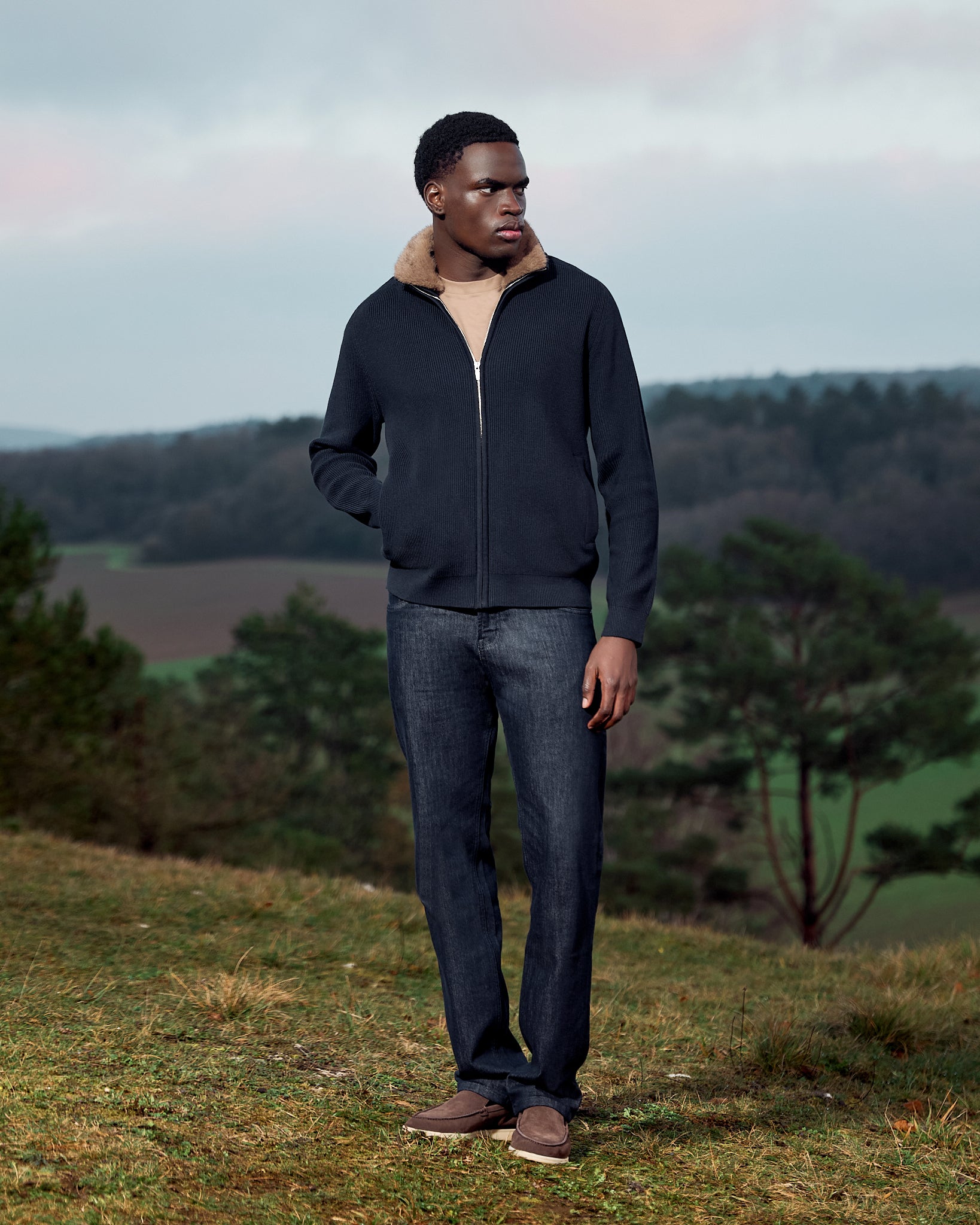 Man wearing a dark jacket and jeans standing in a natural landscape with trees and open sky.
