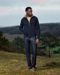 Man wearing a dark jacket and jeans standing in a natural landscape with trees and open sky.