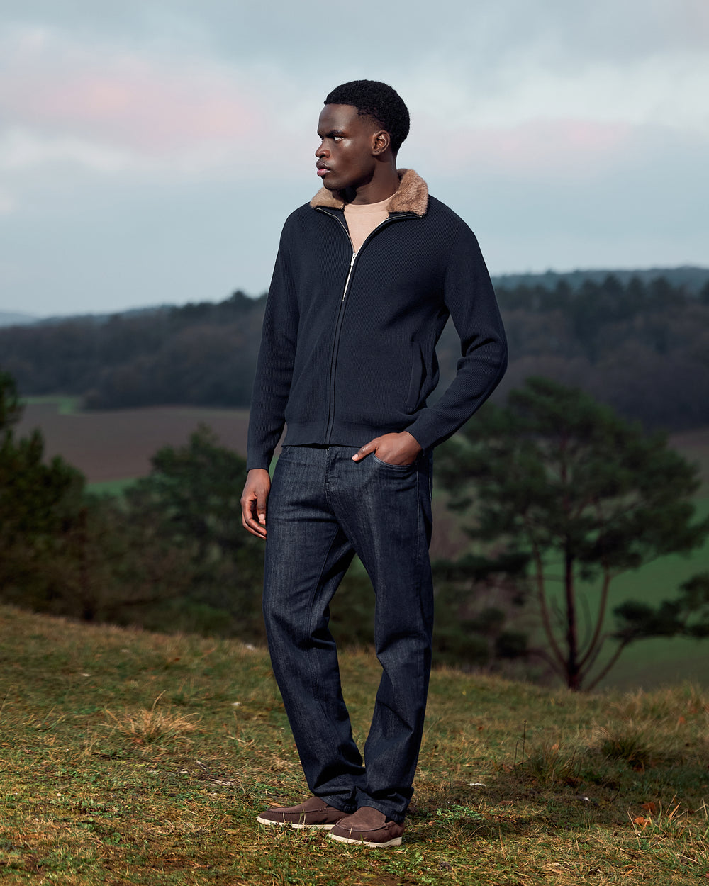 Man wearing a dark jacket and jeans standing in a natural landscape with trees and open sky.