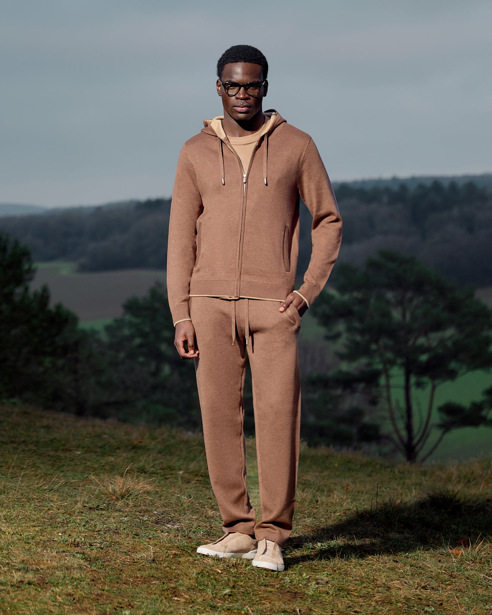 Man wearing a brown tracksuit standing in a field with trees in the background