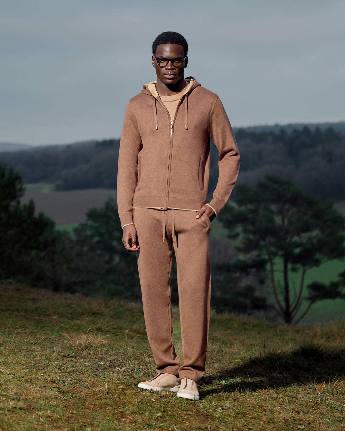Man wearing a brown tracksuit standing in a field with trees in the background