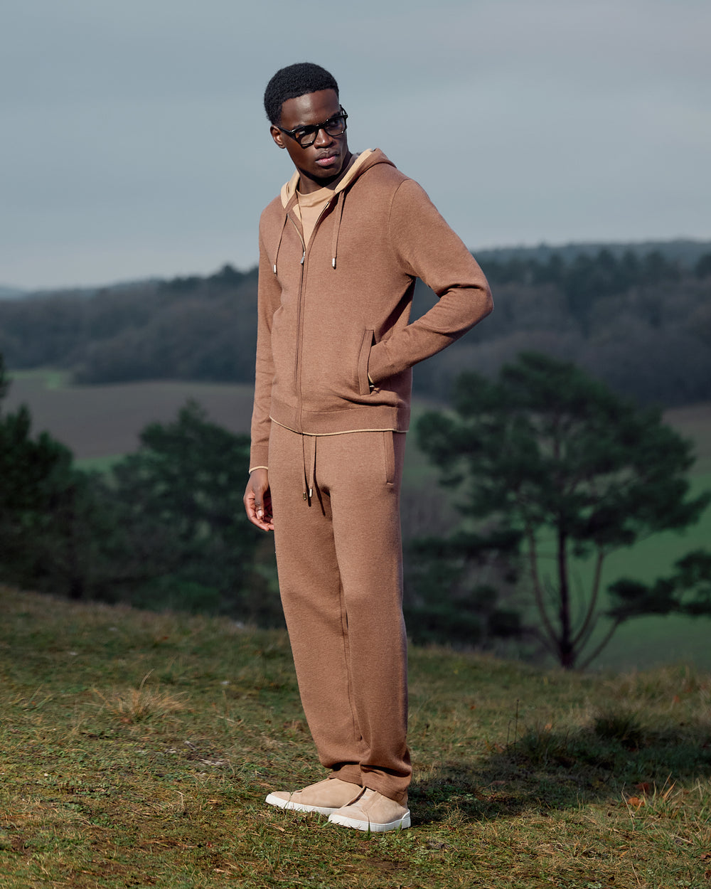 Man wearing a brown tracksuit standing in a natural setting with trees and grass.